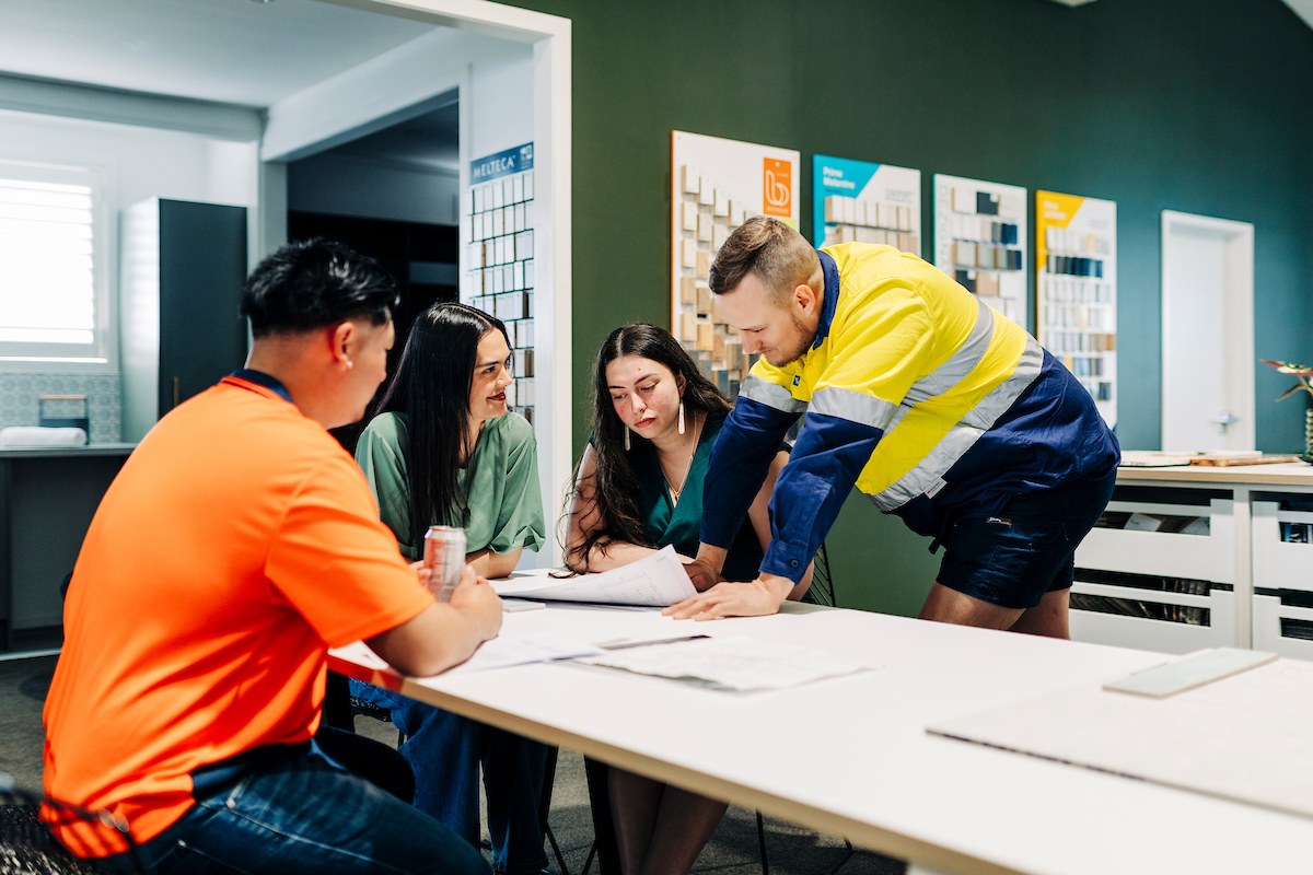 Two tradesmen and two women discuss blueprint plans at a table