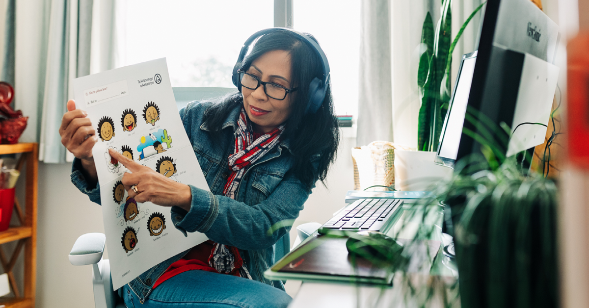 Woman wearing headphones sits at a computer, holding up a Māori language learning poster and pointing to it on camera.