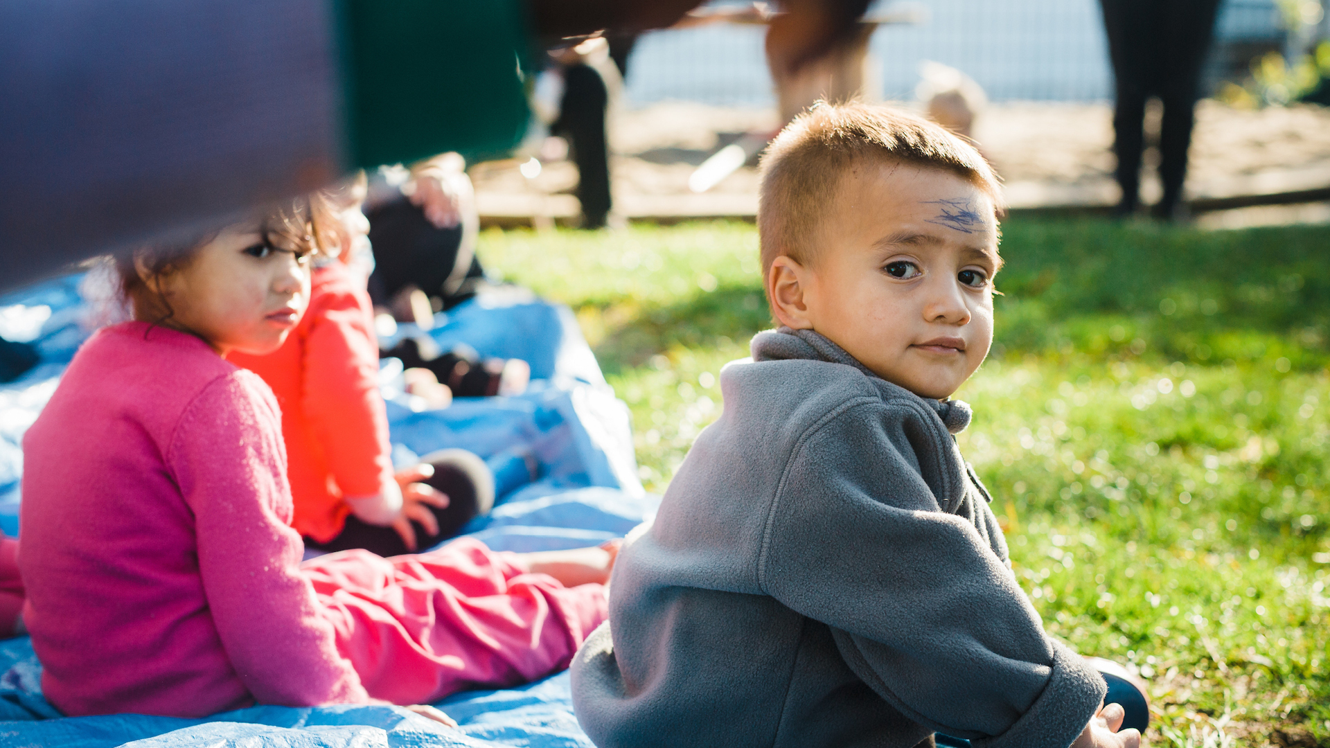Boy looks at camera while children sit on grass outside