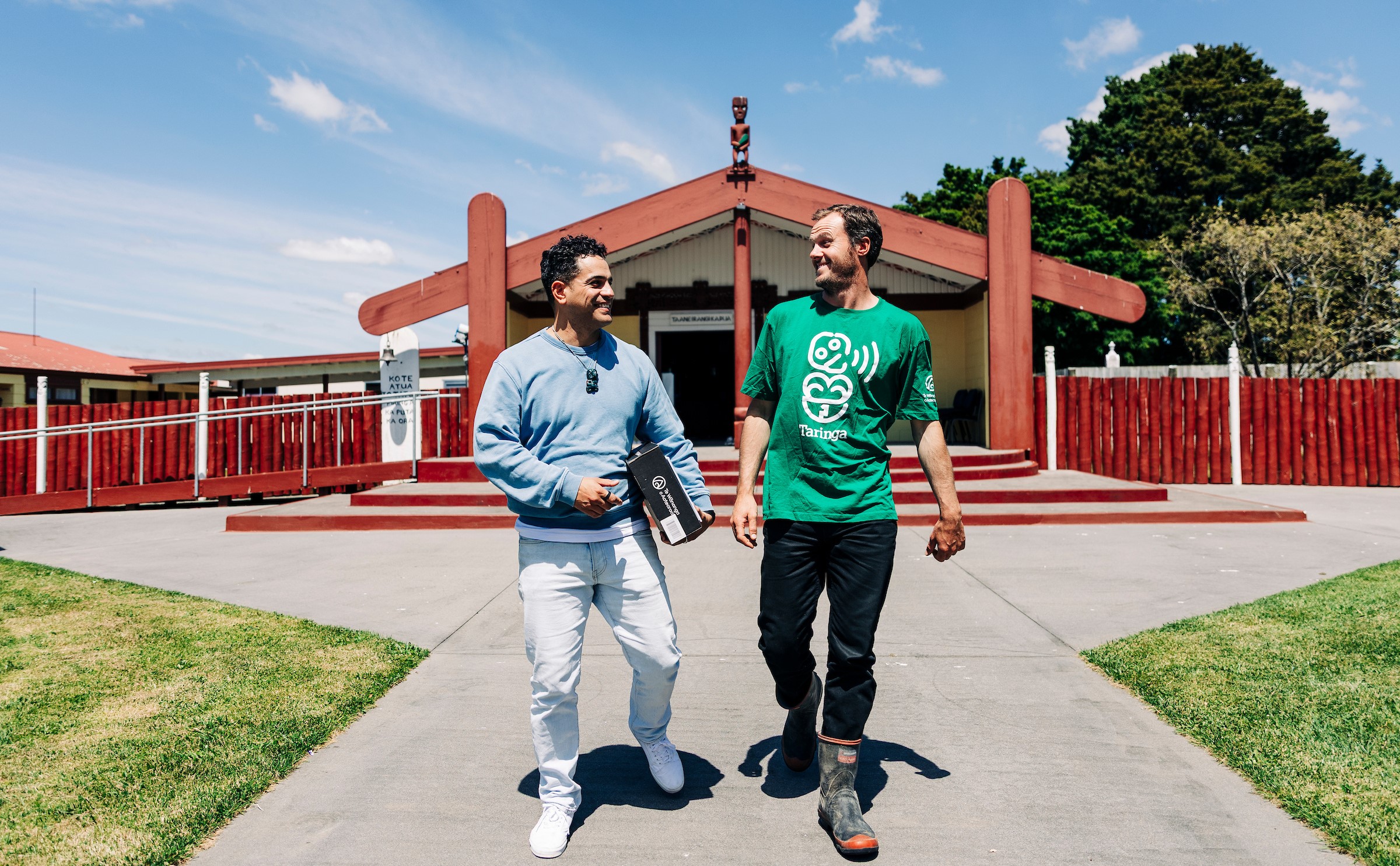 Two men walk outside in front of the wharenui, a traditional Māori meeting house