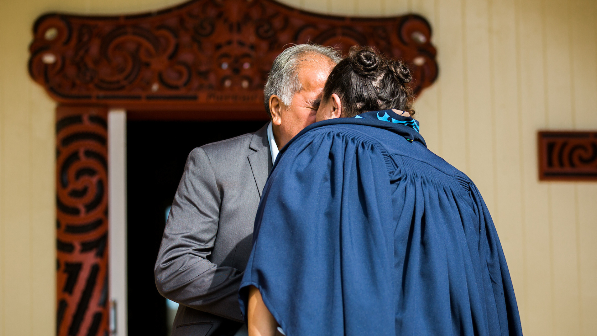A woman in graduation robes and a man hongi outside the wharenui