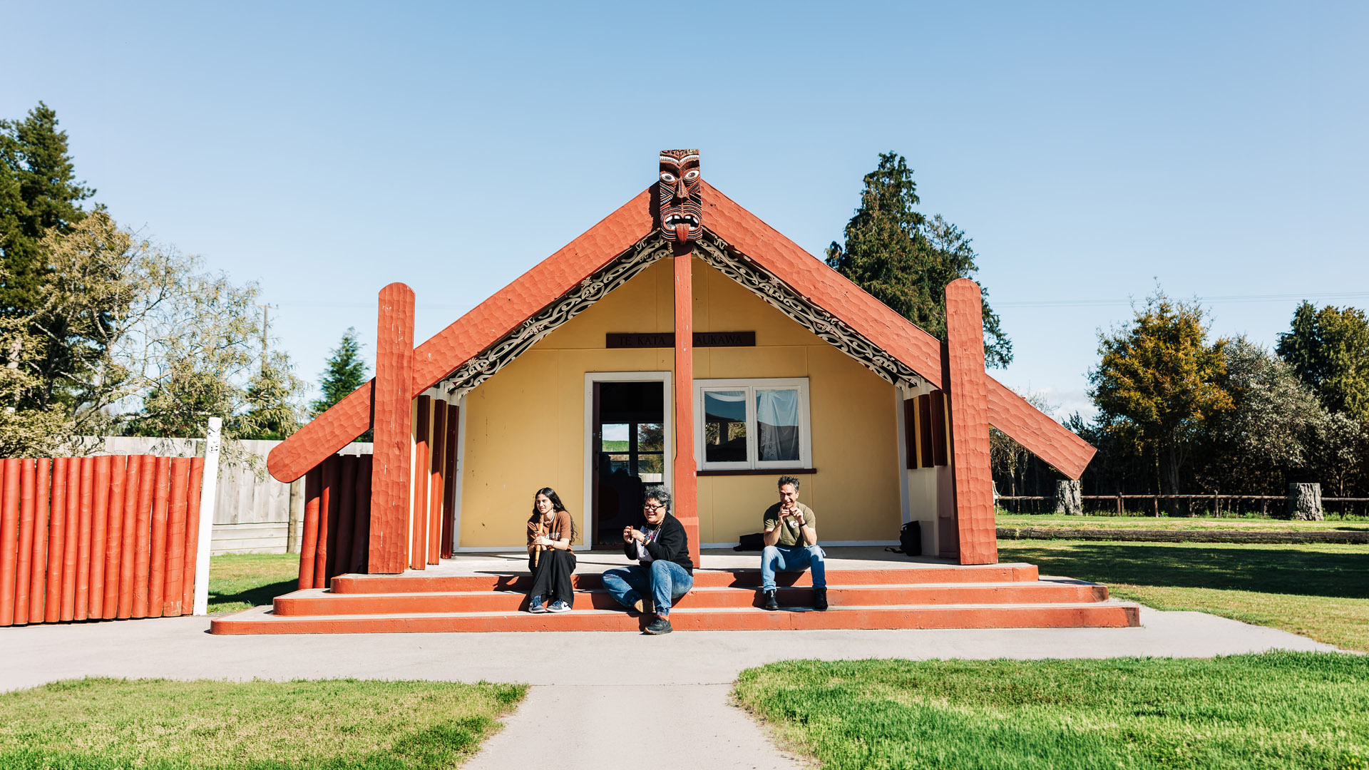 Three people sit on the steps outside a wharenui, each playing taonga puoro traditional Māori instruments