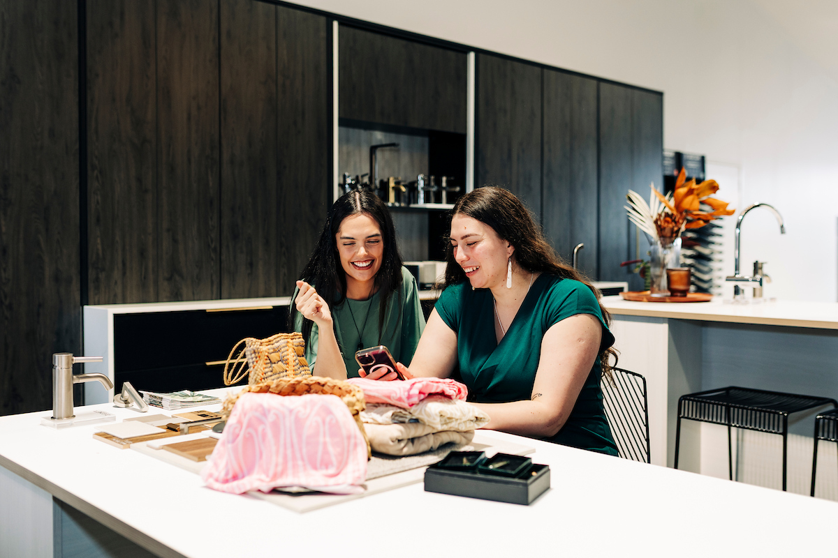 Two women sit at a kitchen table and take photos of Māori homewares