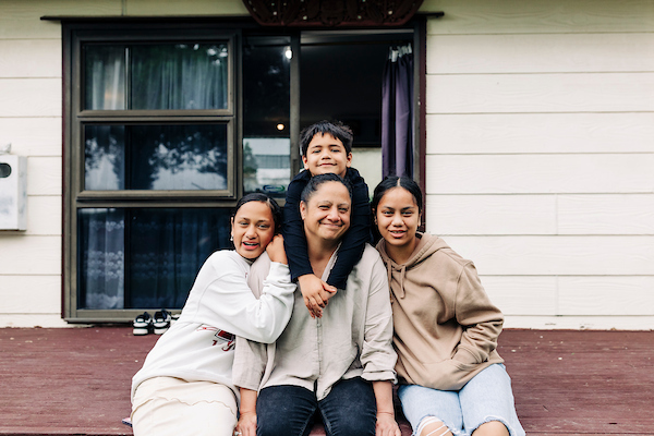 A mum and her kids sit on outside their house on the deck, smiling together