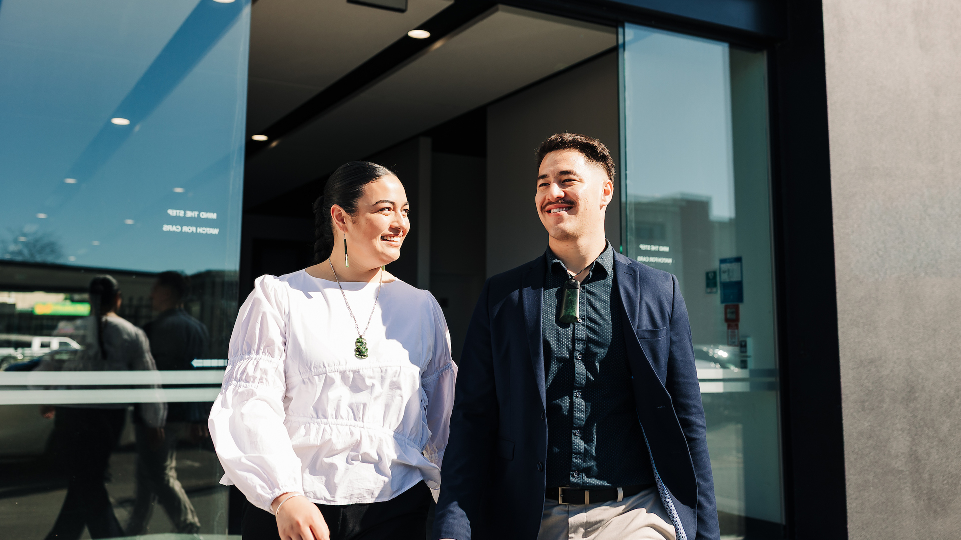 A professional man and women smiling as they walk outside through sliding doors