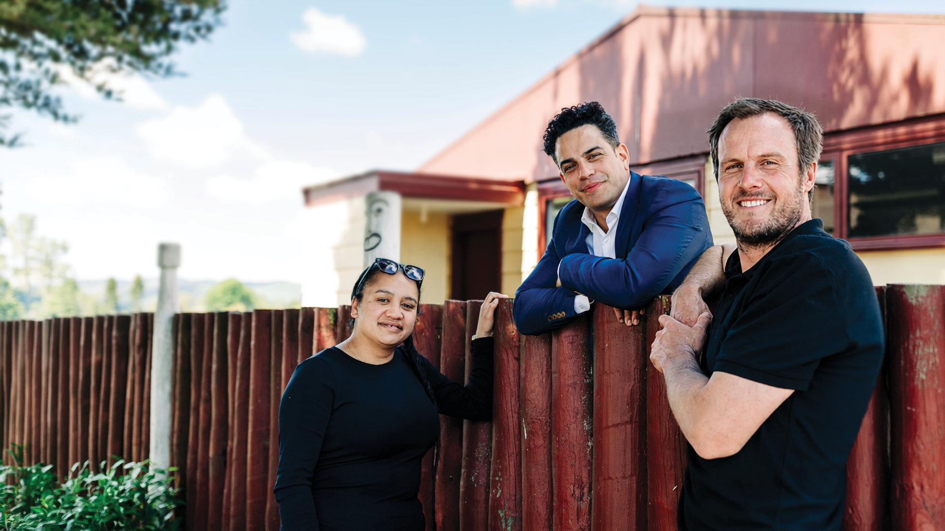 Three adults lean against the fence of a pā as they smile at the camera