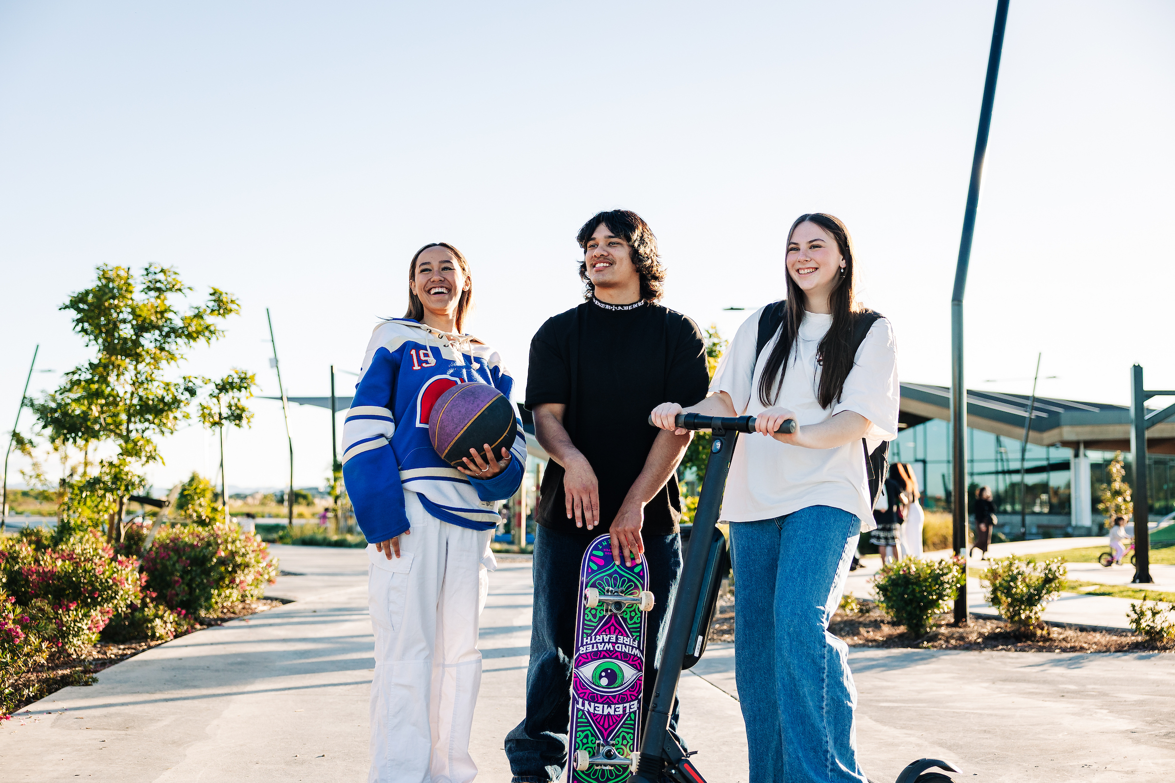 Three young adult students with a scooter, skateboard and basketball in the campus carpark