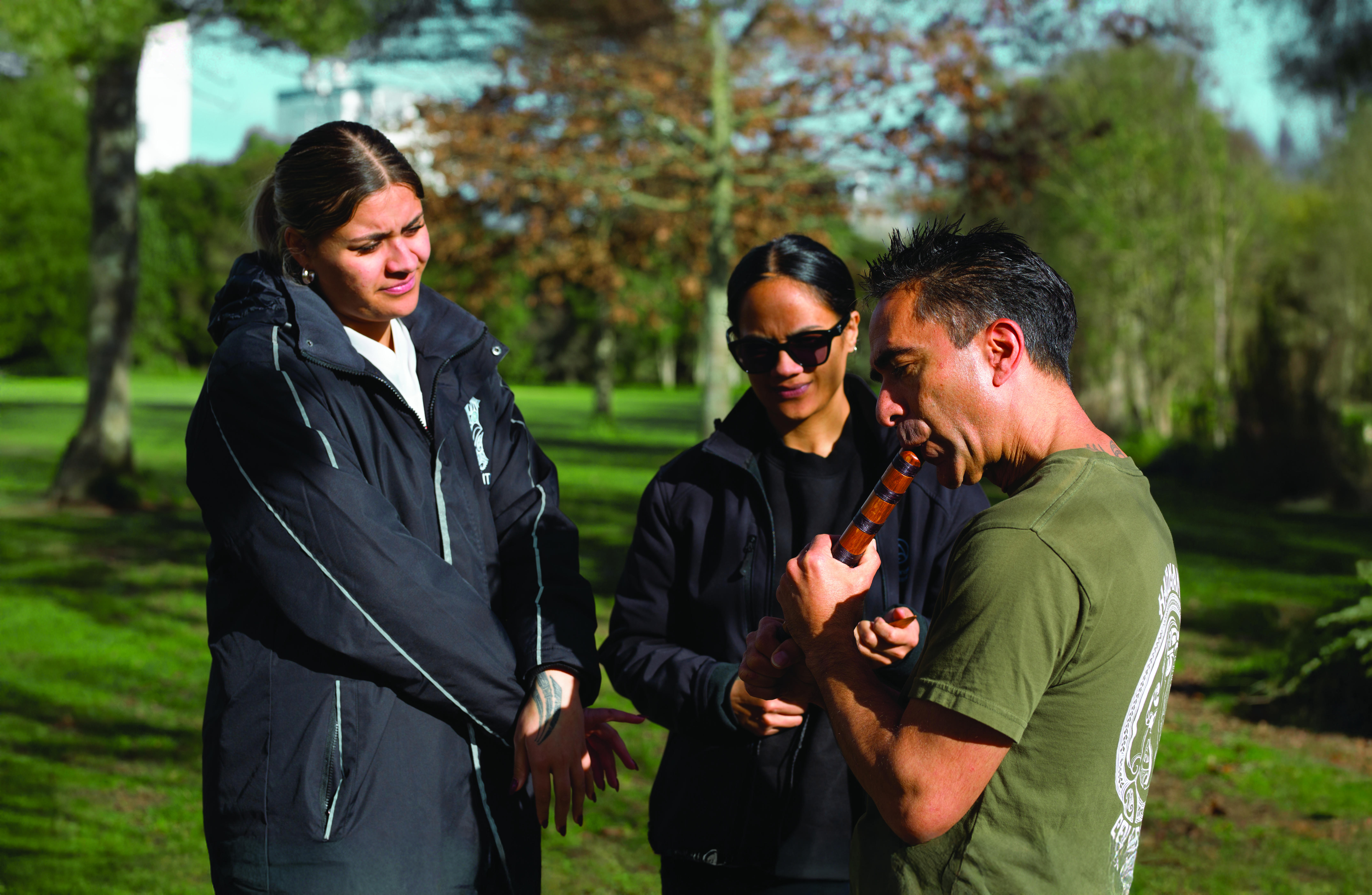 Two women watch a man play a traditional Māori instrument