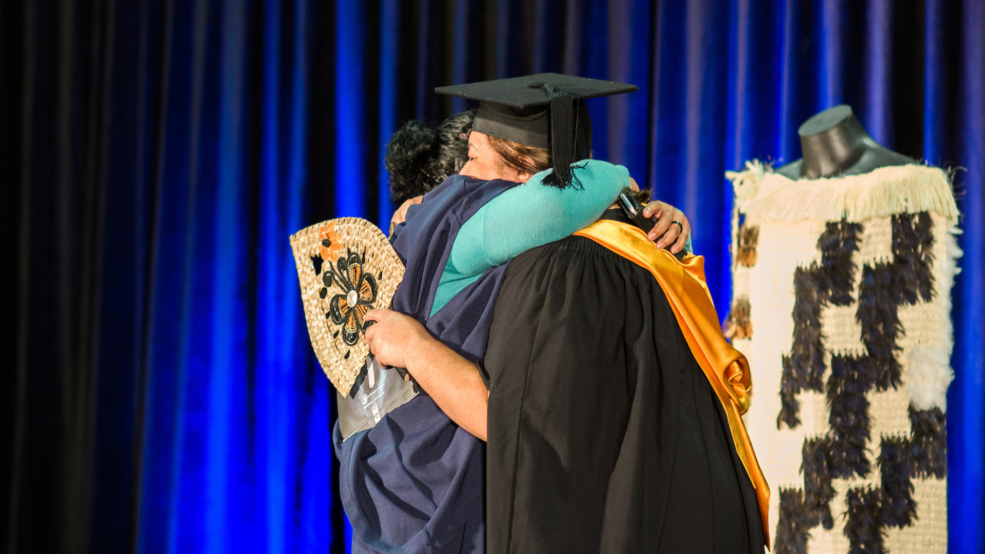 Two people in academic dress embrace on a blue curtained stage